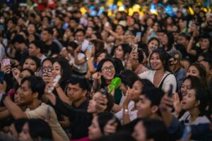 A buzzing crowd of fans, cheering with energy, dressed in vibrant pop culture outfits and waving character merch in the air.